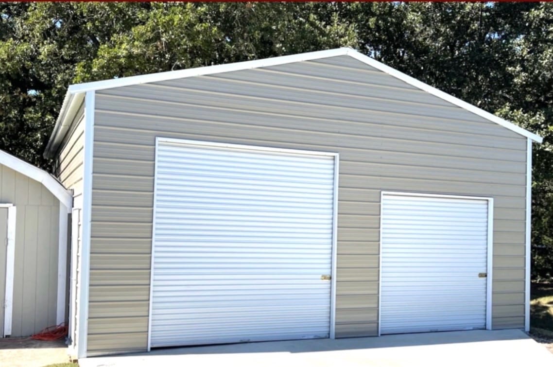 Tan steel garage with a white trimmed gable end, one wide roll-up door and one narrow roll-up door on a concrete slab